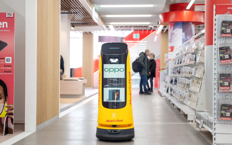 Gelber Werberoboter präsentiert Angebote autonom im Elektronikmarkt zwischen Regalen und Kunden